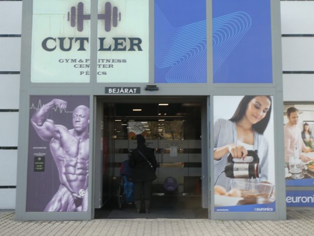 Közelről látjuk a Cutler Gym bejáratát, széles, automata ajtóval. Éppen beléptek Gergő és Evelin az edzőterembe.