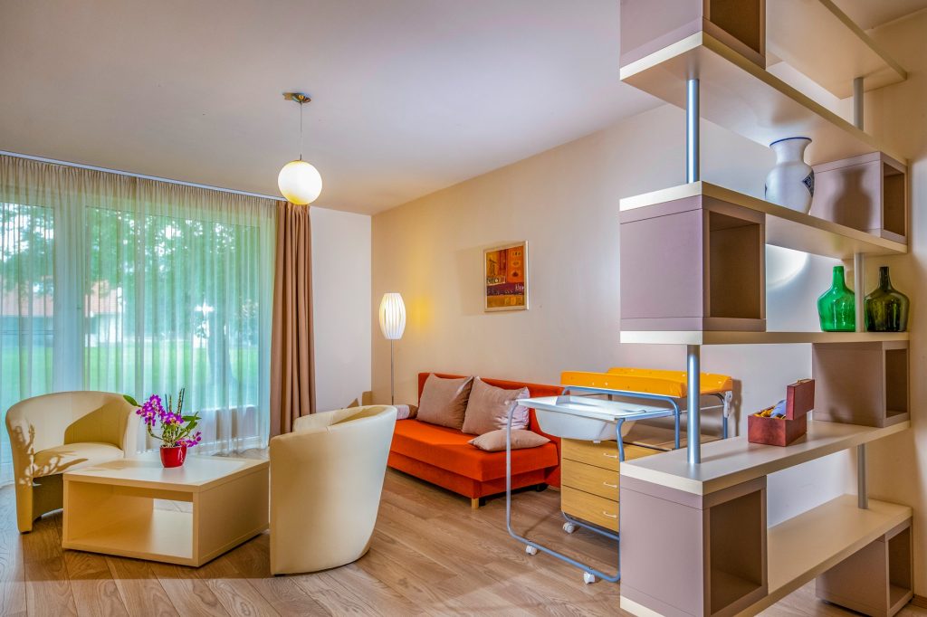 Az akadálymentes apartman nappali részében a sarokban kanapé, a szoba közepén két fotel és egy asztalka látható. A kanapé mellett pelenkázó asztal bekészítve. A térbe benyúlik egy faltól-falig érő polcrendszer. A burkolat parketta.