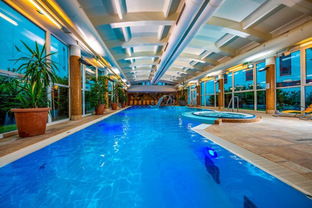 A hotel fürdő részlege látható a képen, feszített víztükrű medencével. A medence partja sima térkő burkolatú. 